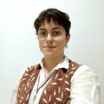 Fotografia de Andreia Duarte Costa, vestida com uma camisa branca e colete castanho com folhas douradas. Possui um colar em forma de folha a condizer com o colete. A pessoa tem cabelo curto castanho e olhos castanhos e encontra-se a sorrir ligeiramente para a foto. Usa também óculos de armaçao dourada e apresenta um brinco na orelha.